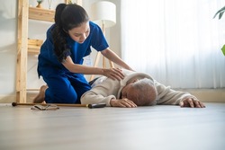 Asian caregiver helping senior male from falling on the ground at home. Elderly older patient having an accident after doing physical therapy then rescued by attractive therapist nurse in living room.