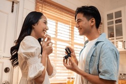 Asian romantic man making surprise proposal of marriage to girlfriend. Attractive young male and proposing to beautiful happy woman, with wedding ring enjoying surprise engagement in kitchen at home.
