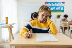 Caucasian young boy student doing an exam test at elementary school. Adorable children sitting indoors on table, feeling upset and depressed while writing notes, learning with teacher at kindergarten.
