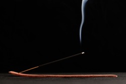 Smoke curls of incense stick in wooden holder for relaxation and meditation exercises, black background. Aromatherapy session with burning aroma stick, pleasant aroma and incense