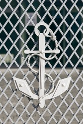 White anchor with mooring rope on mesh fence as symbol of navigation and maritime, white fence in front of entrance to main city port. Decorative navy anchor symbol of marine defense
