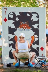 Girl artist draws with paint brush surreal animal playing card on white canvas at outdoor art painting festival, paintings art picture process, rear view. Woman artist paints atmospheric picture