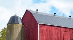 red wooden barn with concrete silo on small hobby farm in Ontario barn with new silver metal roof freshly painted red wood slats horizontal format room for type content or logo blue sky in background 