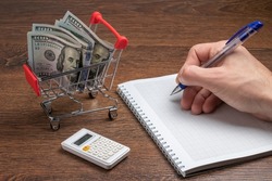 Shopping planning. Man writes a shopping list on a piece of paper. Shopping cart with dollars banknotes and calculator and paper