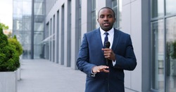 Portrait shot of young African American handsome male journalist talking with microphone for news episode outdoor. Pandemic concept. Man correspondent in suit and tie and with mic.