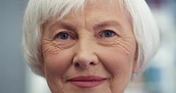 Close up of the Caucasian old happy and cheerful lady with grey short hair looking straight to the camera and smiling. Indoor.