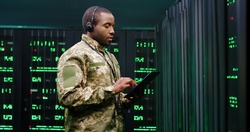 African American military man in uniform and headset working in servers room, tapping on tablet device and speaking in mic. Work with secret information in army. Digital data defence concept.