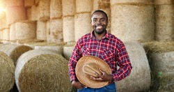 Young handsome African American man farmer standing and smiling in barn with hay barn. Portrait of happy cheerful male shepherd taking off hat and doing greeting gesture in stable. Outdoor.