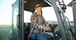 Portrait of beautiful Caucasian young woman in cap sitting in big tractor machine and smiling cheerfully to camera. Pretty happy female farmer worker in field at farm. Agricultural work.