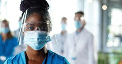 Close up of African American woman physician in face shield looking to camera in clinic. Portrait of female doctor or nurse in medical mask and goggles in hospital. Docs talking on background Covid-19