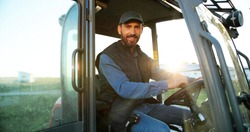 Portrait of young Caucasian male farmer in cap sitting in tractor with open door and smiling to camera. Field farming vehicle. Machine for agriculture. Handsome smiled man.