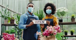 Portrait of joyful African American male and female in masks standing in flower shop and holding Open sign. Woman and man florists workers looking at camera in good mood. Family business concept