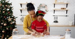 Portrait of happy African American mom with little cute kid in xmas sweater standing at table in home decorated kitchen and smiling to camera while making dough for christmas gingerbread cookies