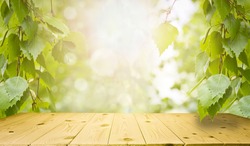 Spring and summer background - fresh green birch leaves, frame in the rays of sunlight, with a wooden table. Abstract natural backgrounds to showcase and promote your product.