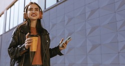A stunning young woman walking down the street with a cup of coffee, against a backdrop of a gray wall. Happy smiling girl listens to music through the headphones and  holds a cellphone in her hand.