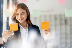 Young smiley attractive, businesswoman using sticky notes in glass wall to writing strategy business plan to development grow to success