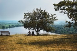 Two people are sitting under a tree on the river bank, a color photo.