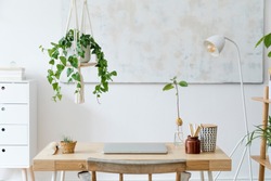 Stylish and boho home interior of open work space with wooden desk, chair, lamp, laptop and white shelf. Design and elegant personal accessories. Botany and minimalistic home decor with plants.