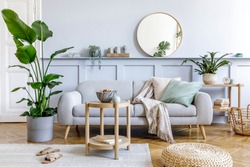 Stylish living room interior with design grey sofa, coffe table, pouf, basket, shelf, mirror, tropical plants, decoration, carpet, pillows and elegant personal accessories in modern home decor.
