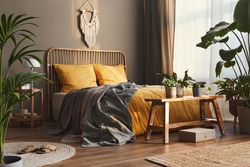 Wooden bed in stylish neutral bedroom interior with design furniture, decoration, carpet, bench, tropical plants, bed sheets, blanket, pillows and elegant personal accessories in home decor.