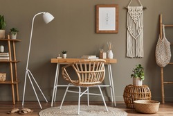 Modern composition at boho interior of home office room with wooden desk, stylish armchair, bamboo shelf, carpet, macrame, mock up poster frame, office supplies, decoration and personal accessories. 