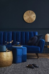 Elegant modern living room interior design with glamour blue velvet sofa, pouf, black metal shelf, poster, plants and modern home accessories. Dark blue wall. Template. Copy space.