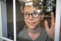 Blond child with glasses, he looks calm and serene through the window with his hand resting on the glass. Lights of the sunset are reflected on the glass. Wait for friends to come play with him