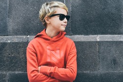 Portrait of a beautiful smiling boy with sunglasses leaning on a black wall. Trendy blond teen with red sweatshirt looking away arms crossed. Serious young male. Youth, relax fashion people concept