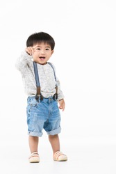 Studio portrait of cute, adorable, Asian toddler boy wearing denim overalls, long sleeve T-shirt, orange shoes, standing, smiling broadly, on isolated white background