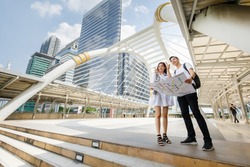 Wide-angle portrait of cute smiling young Asian lover tourists standing and holding paper metro map on footbridge and point to direction together to destination with tall building and sky background