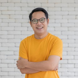 Portrait of adult Asian man wearing  yellow t-shirt and eyeglasses crossed arms and smiling looking to camera with friendly and self confident.
