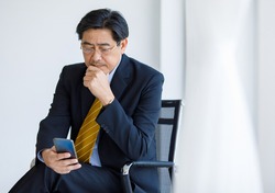 Portrait shot of Asian senior old stress businessman in formal suit and golden eyeglasses sit on chair worrying reading business text message from customer through mobile smartphone in company office.