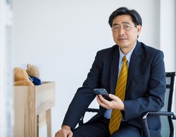 Portrait shot of Asian senior old stress businessman in formal suit and golden eyeglasses sit on chair worrying reading business text message from customer through mobile smartphone in company office.