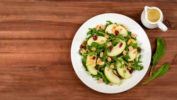 Green apple slice salad with rocket, oat granular, dried cranberry, and cashew nut placed in white dish on wooded table. Oil vinegar salad dressing, a side dish. Two rocket leaf used as decoration.