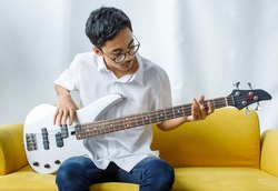 Portrait shot of handsome smiling young teenager enjoying playing the bass guitar. Junior guitarist in casual clothes sitting on the yellow couch and holding an instrument while looking at camera