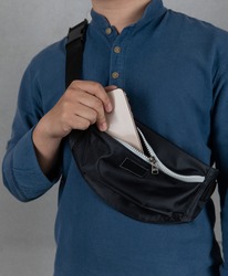 Closeup studio shot of male model in blue long sleeve shirt putting smartphone into front zipper pocket of trendy urban small black crossbody strap casual fanny pack bag in front gray background.