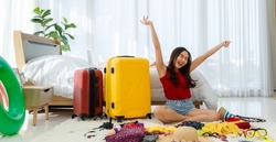 Young happy and exciting Asian female traveler getting ready for a summer holiday trip vacation with her bright yellow suitcase and clothes swimsuit ready to be packed. Throwing hat in the air.