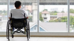 Back view of young Asian handicapped teenage boy without legs sitting on wheel chair and looking to outside of home with copy space on right.