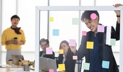 Asian trans man smile and happy at LGBT conference as enjoy explaining idea for business strategy by sticking colorful paper note on glass board while transgender colleagues looking with interesting