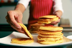 Closeup shot of heap of newly baked yummy fluffy hot golden good smell pancake snack in hand of chef who wears red apron while picking up bread from stack in blurred foreground.