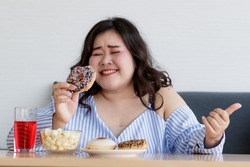 Happiness closeup shot of Asian young happy beautiful hungry overweight fat chubby female smile  hold chocolate donut in hand with dessert cake popcorn on table with happy and enjoy face.