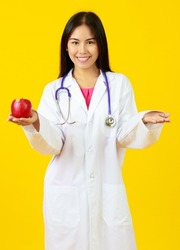 Asian young kind female doctor wearing white gown uniform with stethoscope, happily smiling and holding red apple, presenting nutrition, vitamin, healthy with isolated yellow background
