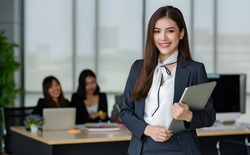 Portrait of young attractive Asian female office worker in formal business suits  smiling at camera in office with blurry colleagues sitting in office as background.