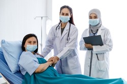 Portrait shot of two female Muslim and Asian doctors in white lab coat uniform with stethoscope standing holding senior patient hand and clipboard smiling under face mask look at camera together.