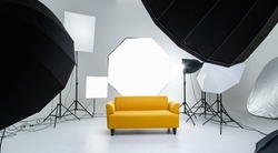 Studio shot fashion backstage photographing shooting set with yellow cozy sofa couch and photography equipment softbox flash strobe spotlight tripods reflector umbrella on white backdrop background.