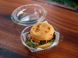 Closeup shot of take away delicious tasty yummy homemade juicy patty grilled beef cheeseburger with vegetable and melting thousand island sauce in disposable plastic round box package on wood table.