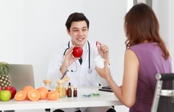 Asian male professional nutrition science dietitian doctor in white lab coat and stethoscope holding red apple and supplement pill explaining discussing weight loss decision choice to female patient.