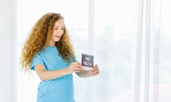 Caucasian young beautiful long curly hairstyle healthy pregnant mother in casual outfit standing holding showing baby ultrasound Xray film picture sonogram in hands in front of curtain background.