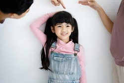 Close up shot of Asian small cute pigtail hair style preschooler child daughter in jeans overalls outfit standing smiling leaning at wall while father using hand measuring checking growth rate height.
