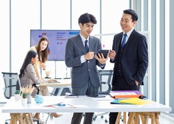 Asian male professional businessmen colleague in formal business suit take coffee break standing holding disposable paper cup and tablet computer talking have conversation together in front monitor.